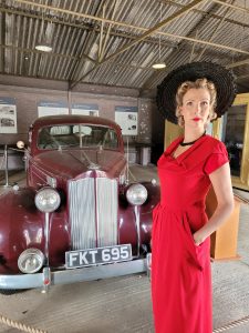 Jayne Darling in red dress and black sun hat standing in front of a classic car