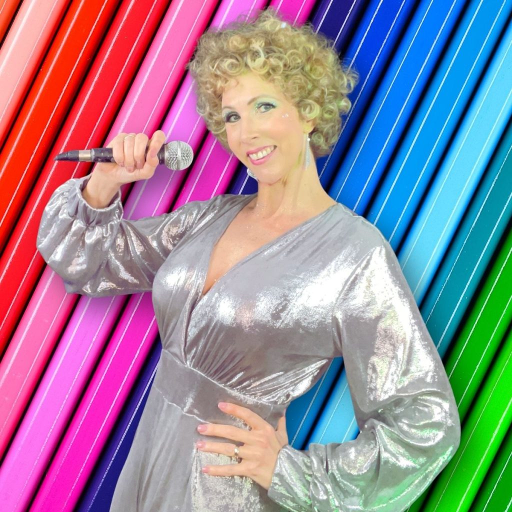 Jayne Darling wears a silver jumpsuit in front of a rainbow backdrop