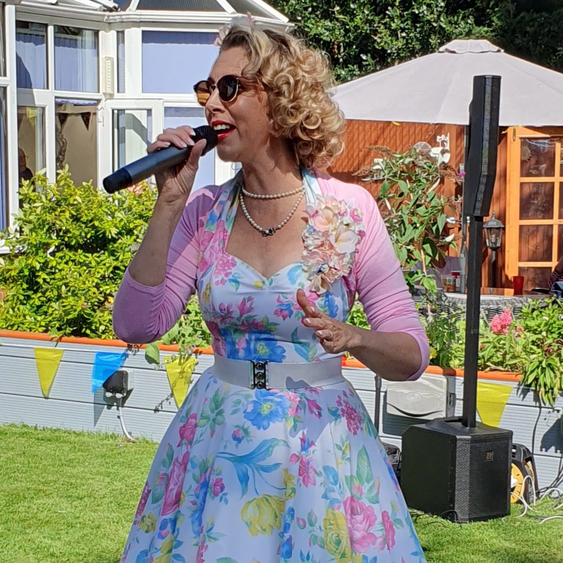 Jayne Darling singing at. garden party using a handheld microphone and wearing a pastel 1950s style dress