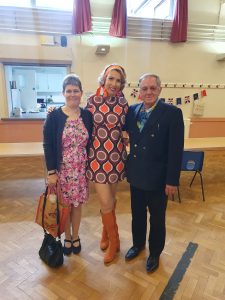 Jayne darling in orange knee high boots in between a man in a suit and a lady in a floral floral dress holding a handbag