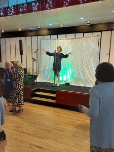 Jayne Darling performs her 1960s show at Clipstone Welfare. She is wearing a black sequin dress and white knee high boots. She is on stage in front of a white curtain