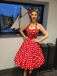 Jayne Darling wears a red polka dot dress and stands in front of a window