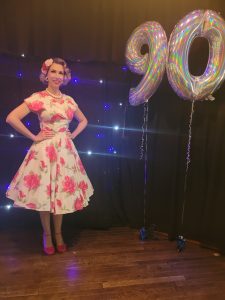 Jayne Darling is shown next to a balloon display. The is wearing a pink floral dress and pink shoes