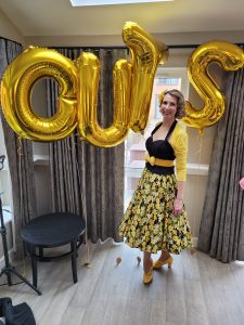 Jayne Darling is shown by some gold balloons , she is wearing a 1950s yellow and black outfit.