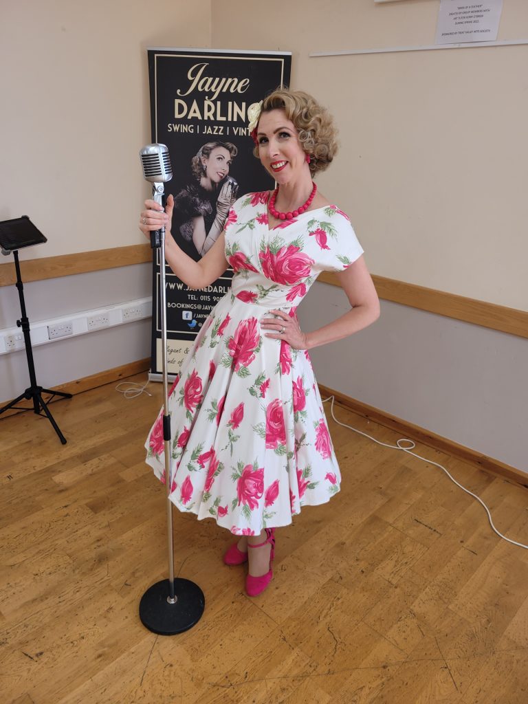 Jayne Darling stands in front of a vintage style microphone , she is wearing a pink floral dress and pink shoes
