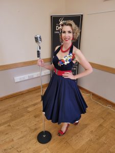 Jayne Darling stands in front of a vintage style microphone . She is wearing a navy 1950s stye dress with red shoes and has flowers in her hair