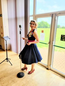 Jayne Darling stands in front of a clear door with a vintage style microphone . She is wearing a navy 1950s stye dress with red shoes and has flowers in her hair