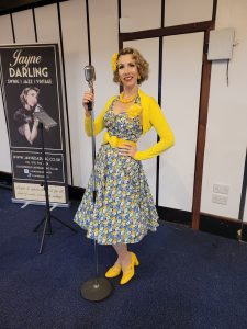 Jayne Darling performs on stage at Clipston Welfare. She is wearing a blue floral 1950s dress and is holding a vintage style microphone