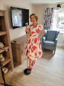 Jayne Darling wears a pink floral dress, holds a vintage style microphone. She is standing in the room of a care home