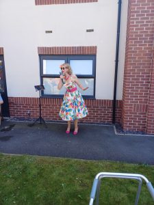 Jayne Darling sings outside a care home wearing a floral Vivien of Holloway dress pink belt and pink shoes . She is holding a vintage style microphone