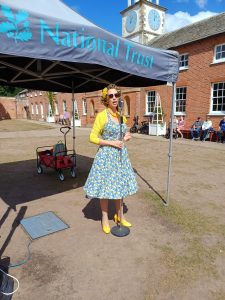 Jayne Darling sings in the sunshine in the courtyard at Clumber park, She is wearing a blue floral dress and yellow cardigan with yellow shoes