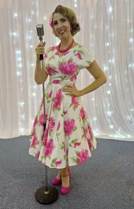 Jayne Darling wears a Vivien of Holloway pink floral dress and is holding a vintage style microphone