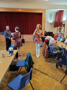 Jayne Darling performs her 1960s. in Sheffield. People dancing and Jayne is wearing white knee high boots, white tights and an orange dress. She is singing into a vintage style microphone