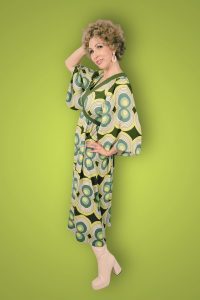 Jayne Darling dressed in a green and yellow retro 1970s dress with white platform boots in front of a green back drop