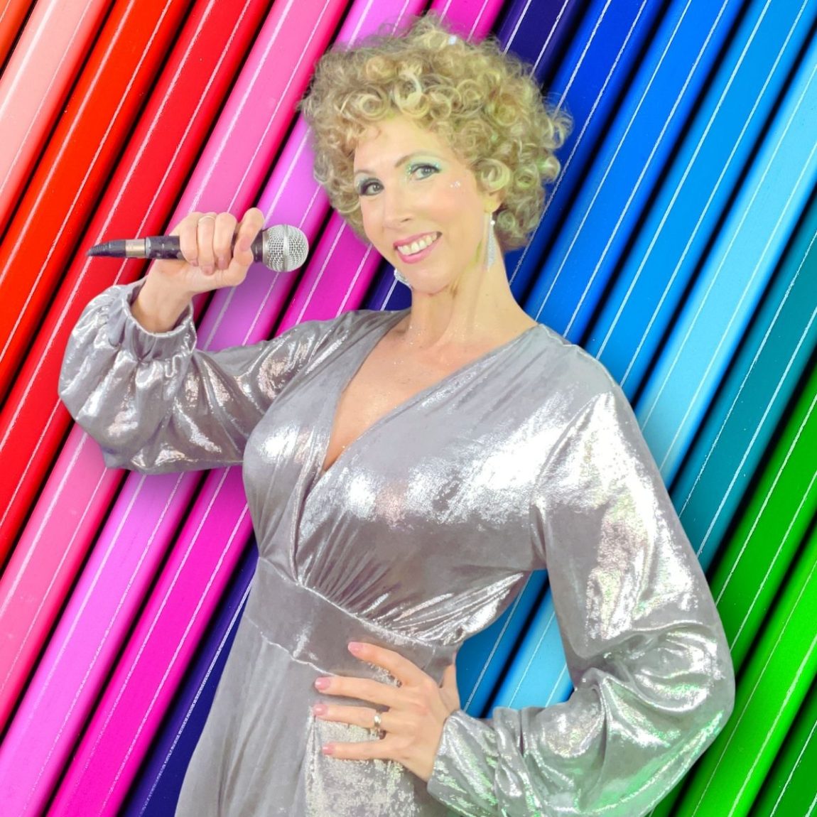 Jayne Darling wears a silver jumpsuit in front of a rainbow backdrop