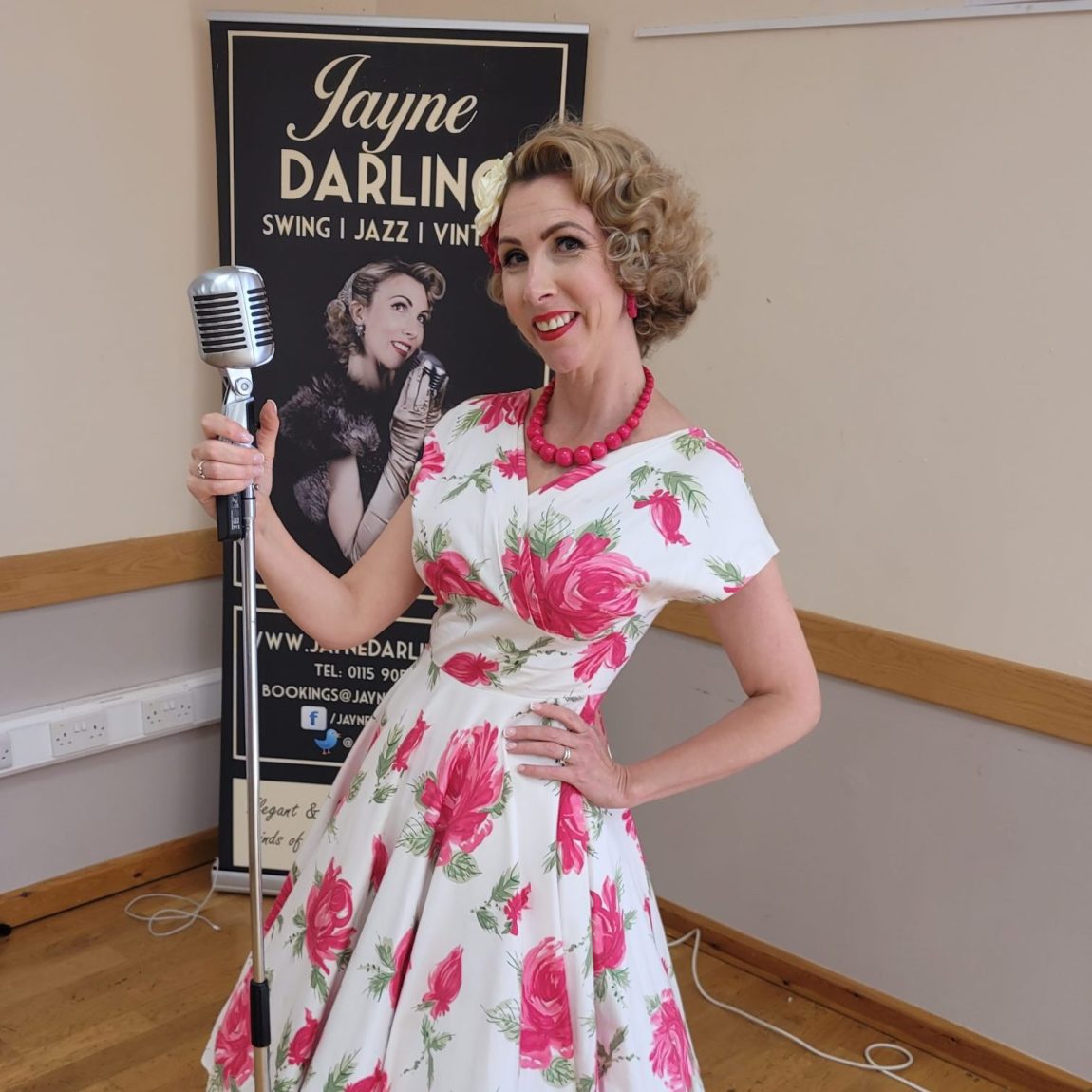 Jayne Darling stands in front of a vintage style microphone , she is wearing a pink floral dress and pink shoes