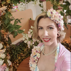 Jayne Darling wearing flowers in her her and as a corsage with a pearl necklace and pink dress stood in front of a large floral wreath